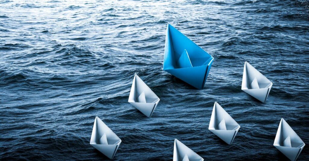 The 4 R's of the Leadership Journey - Risk, Rejection, resilence, Reinvent. Blue paper boat leading the grey boats in water